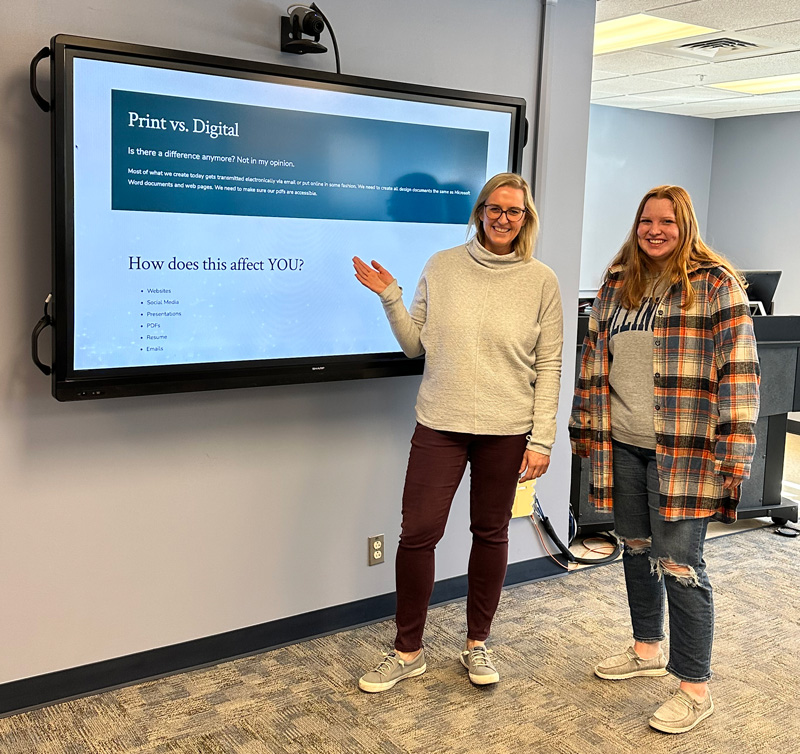 Melissa standing with a student beside a classroom display showing a presentation about print versus digital communication.