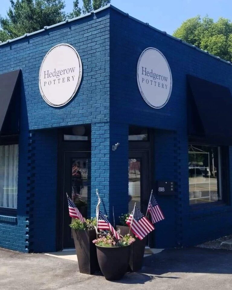 The Hedgerow Pottery storefront, a blue brick building with two round signs carrying the rebuilt logo mounted on two faces above the entrance and the corner window.