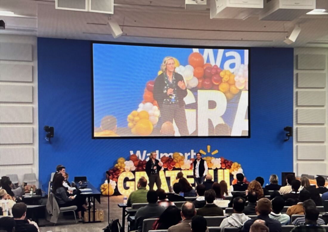 Melissa co-presenting on stage at a Walmart enterprise town hall, her image projected on a large screen above a seated audience.